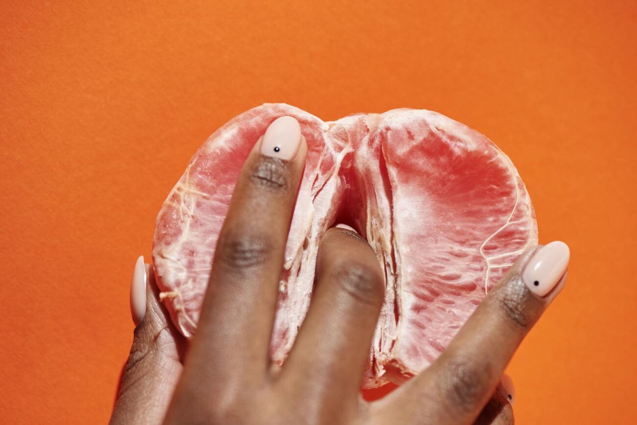A close-up of a hand with polished nails holding a peeled grapefruit against an orange backdrop.