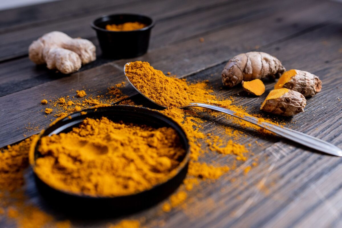 Turmeric powder in a bowl beside fresh turmeric root and a spoon on a dark wooden table